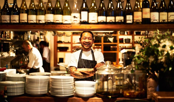 New Cozy Winter Restaurants NYC - A chef at HOUSE BROOKLYN stands with his arms crossed in the middle of the open concept kitchen. The kitchen opening is framed with wine bottles, plates, and other items.